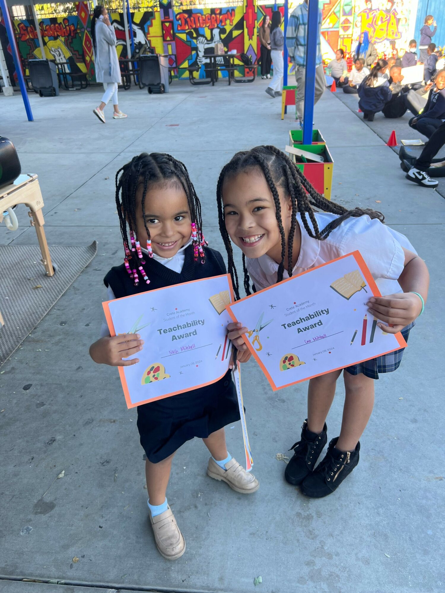 Two young girls holding certificates, smiling, outdoors with colorful murals and other children in the background.
