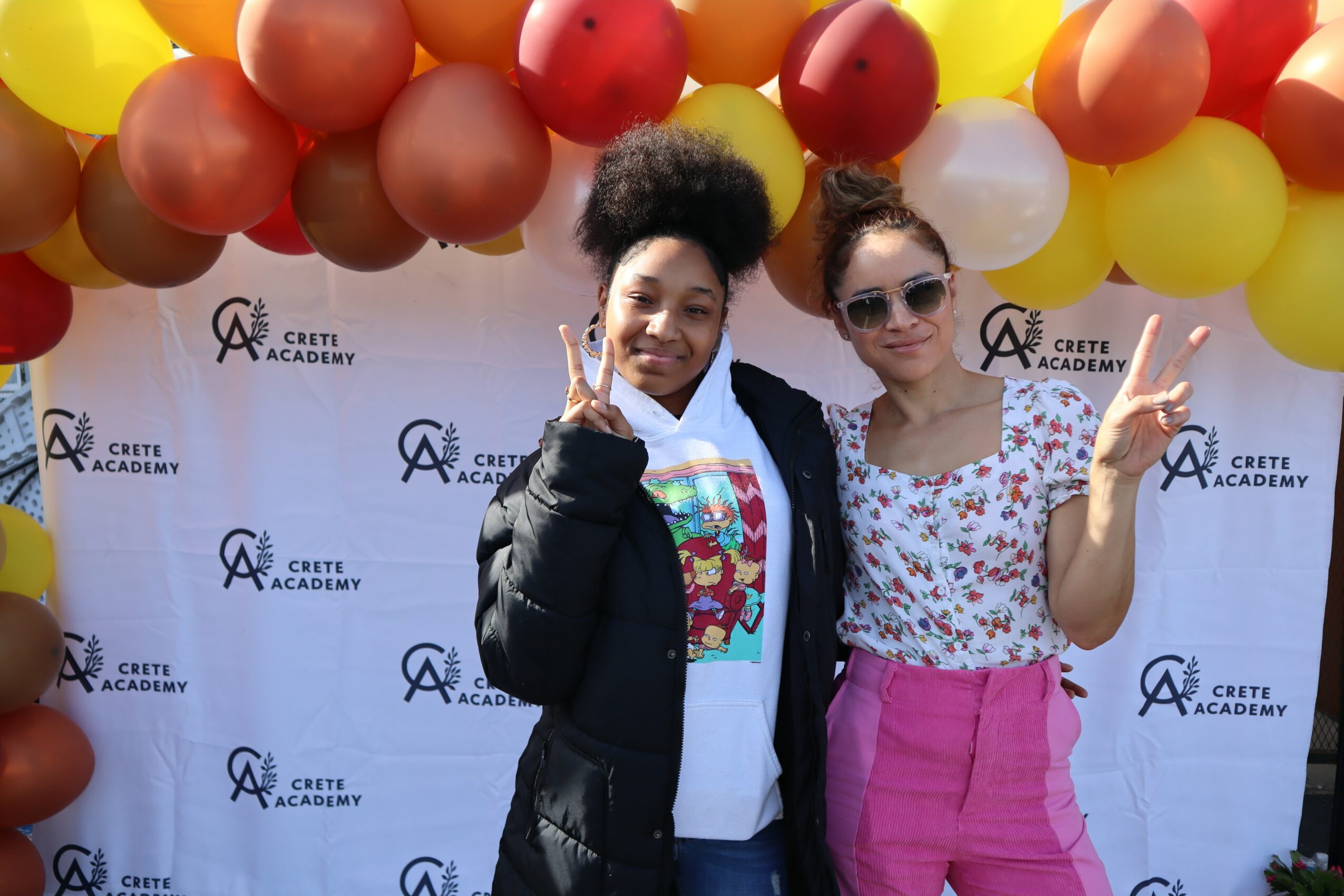 Two women standing in front of a balloon arch and a backdrop with Crete Academy logo, smiling and making peace signs.