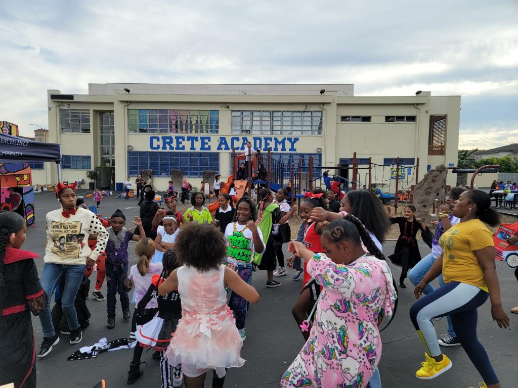 Children and adults gather outside Crete Academy, some dancing and holding signs, with a building and cloudy sky in background.