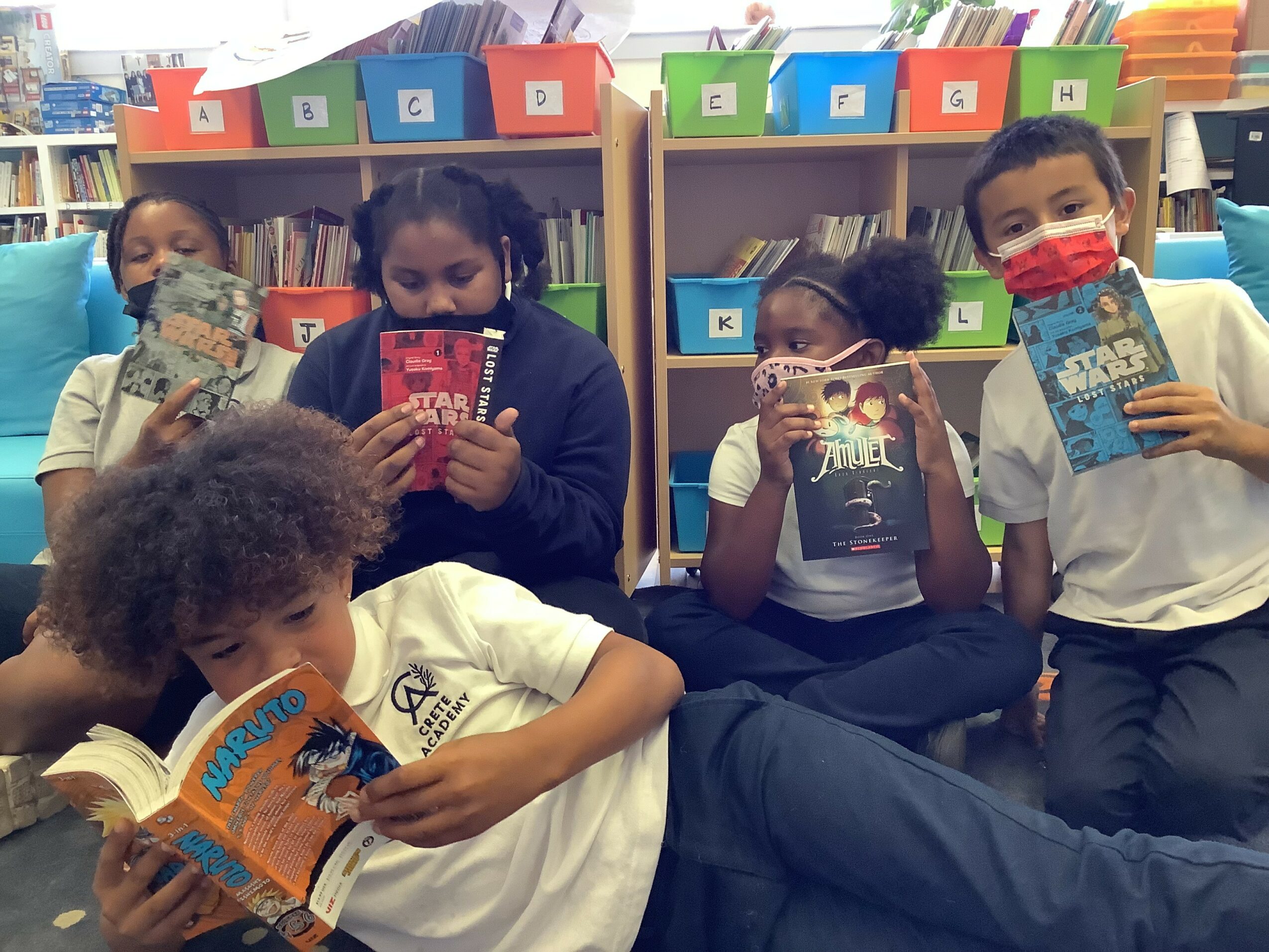 Five children sitting on the floor reading books in a classroom with colorful storage bins behind them.