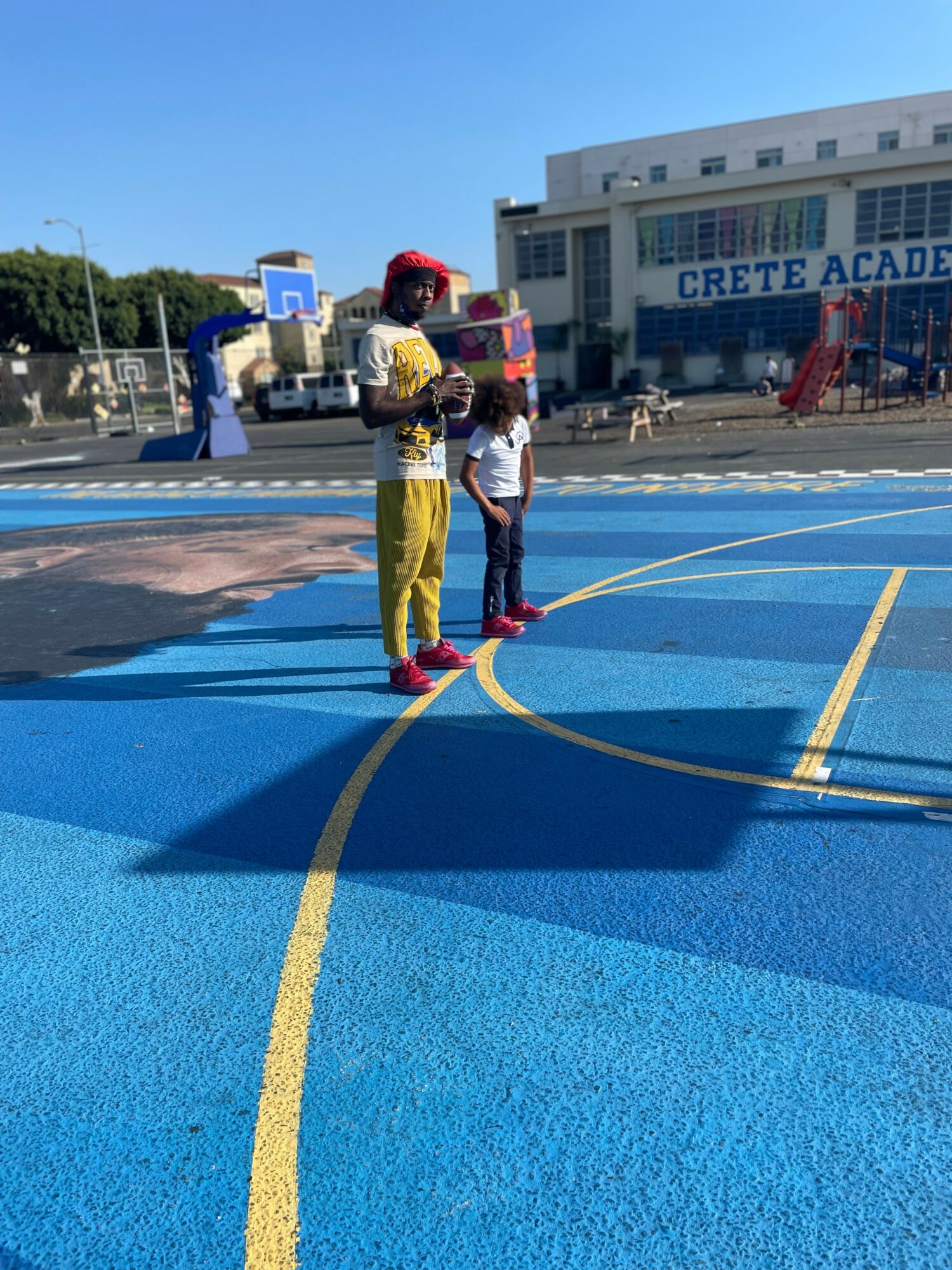 Child and adult standing on a blue outdoor sports court with yellow lines, near a school building and playground.