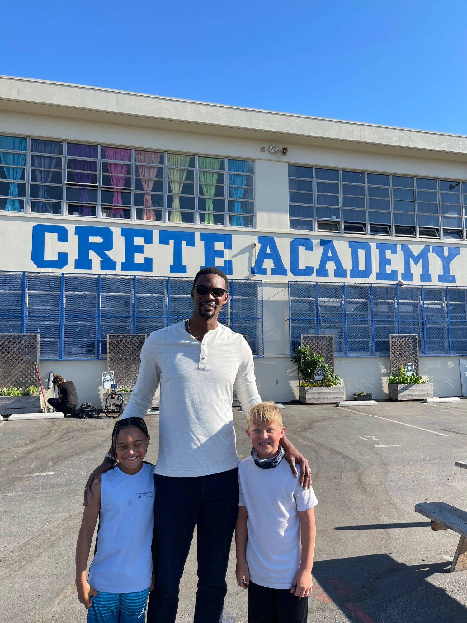 Three people standing in front of a building with a sign reading 'CRETE ACADEMY'.