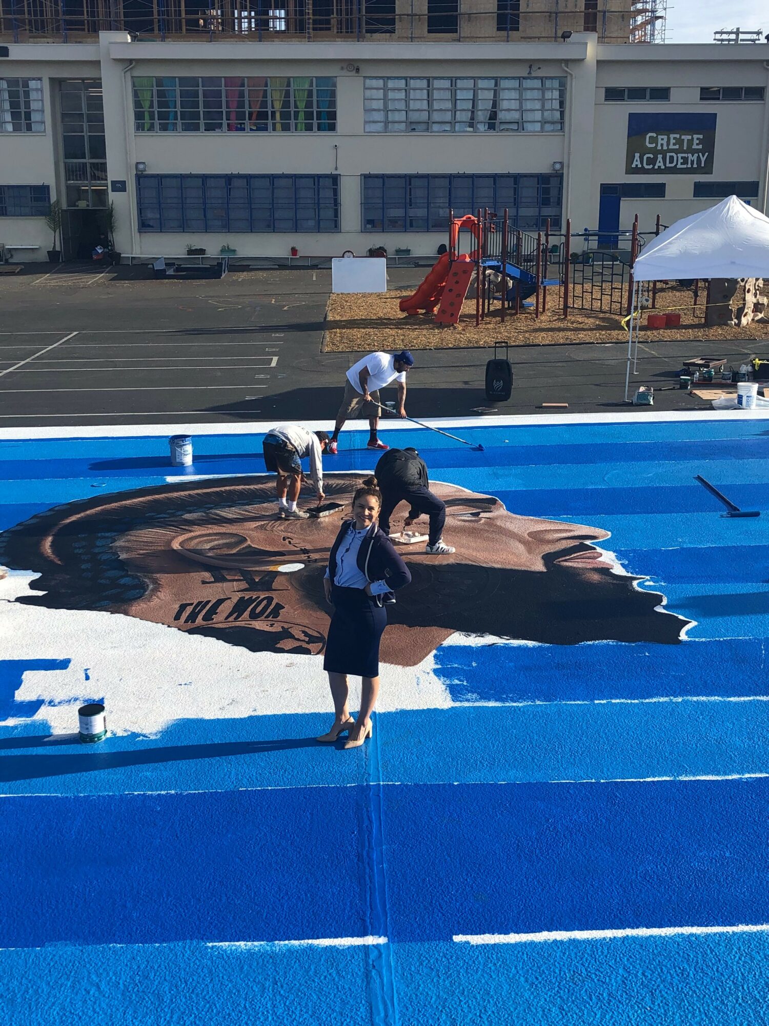 People painting a large portrait of a person's face on a blue surface outdoors, with a school building and playground in background.