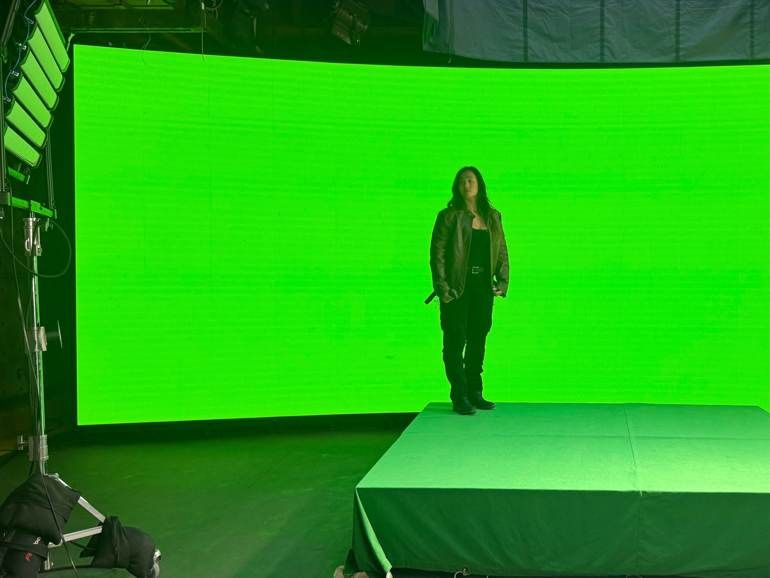 Person standing on a stage in front of a large green screen, wearing dark clothing.