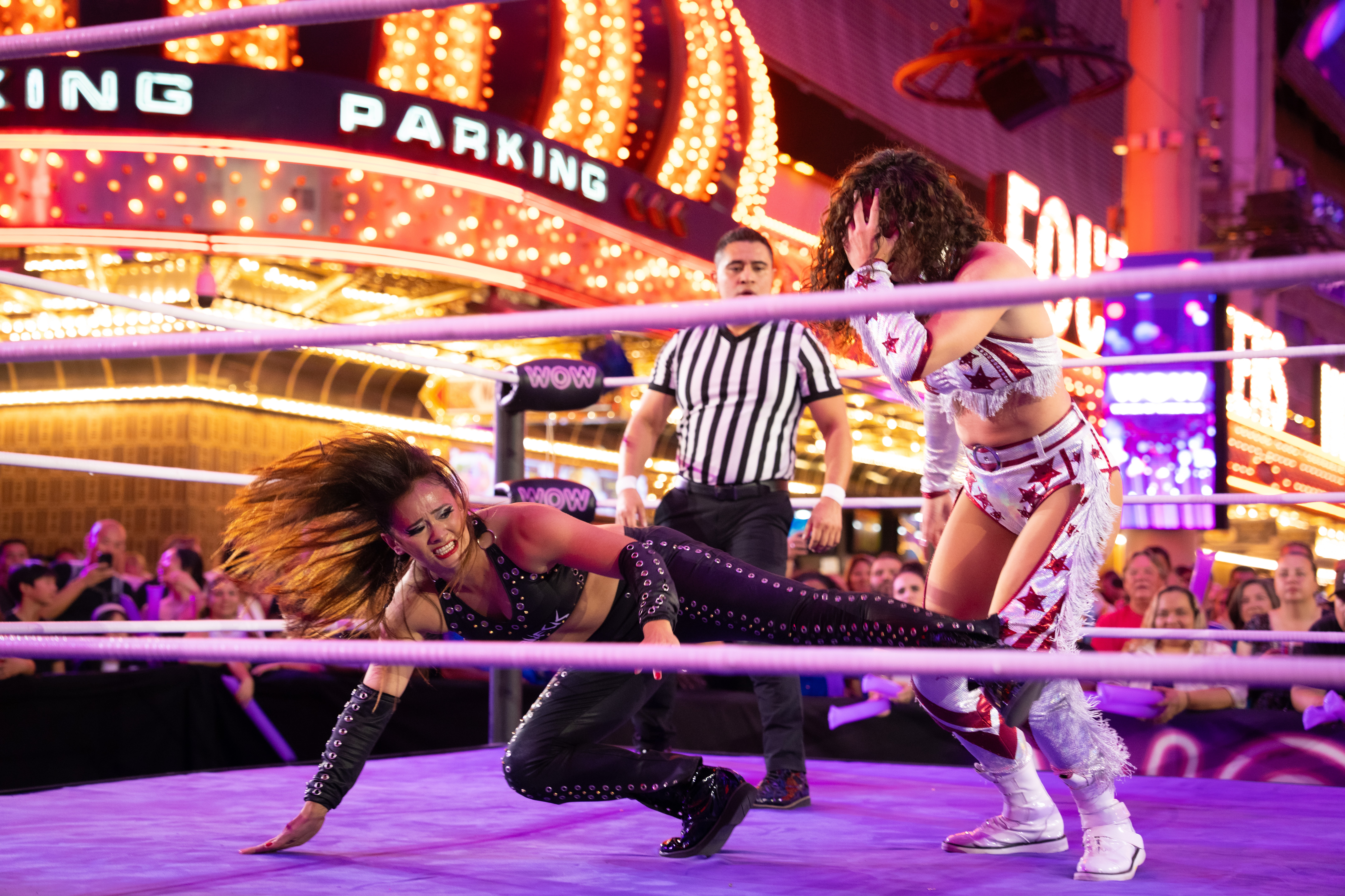 Two women wrestling inside a ring with a crowd and bright lights in the background.