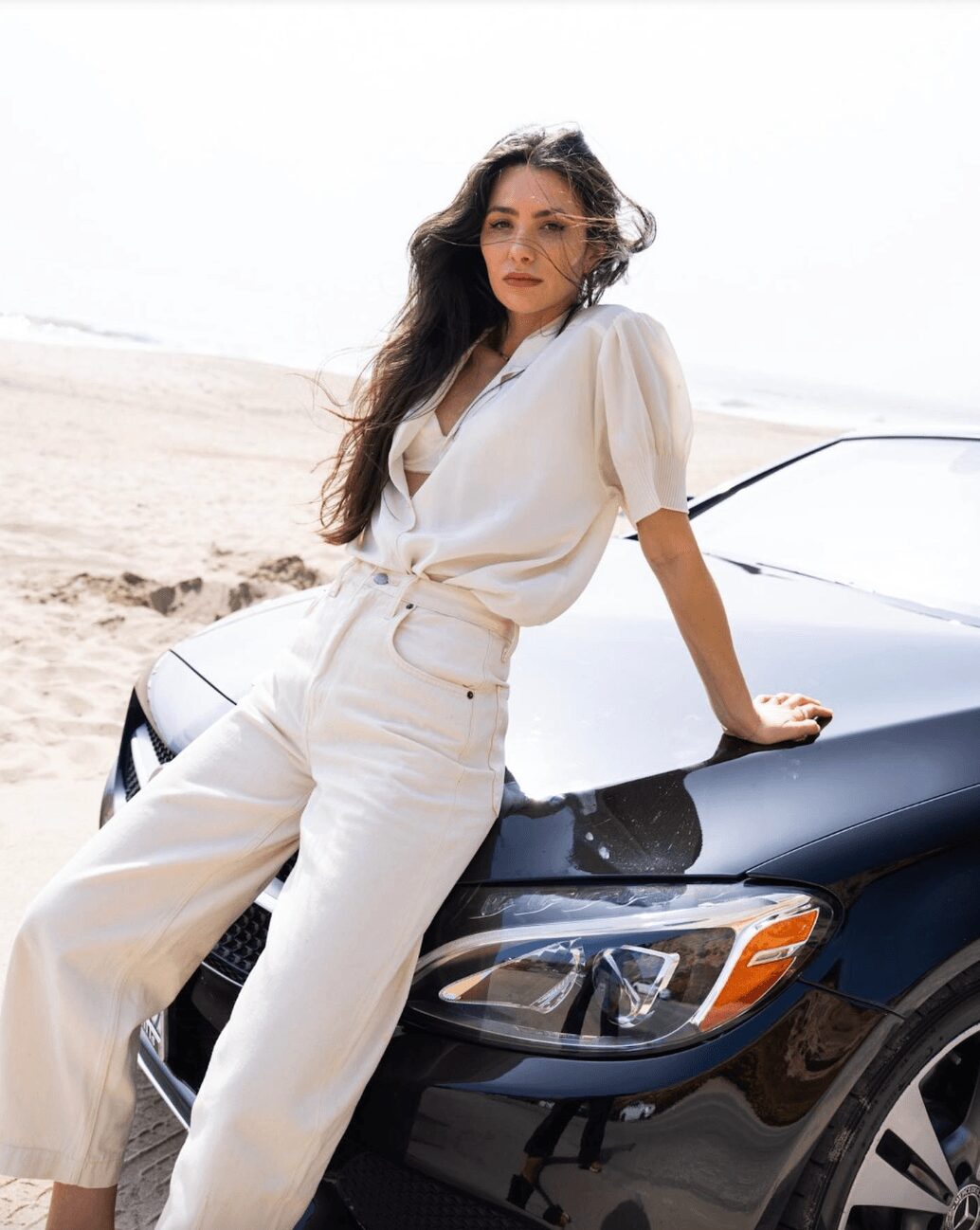 Woman leaning on a black car on a beach, wearing a white outfit, with wind-blown hair and a neutral expression.