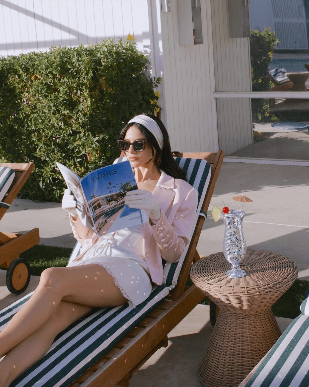 Woman in sunglasses and a hat reading a magazine while lounging outdoors on a striped chair.