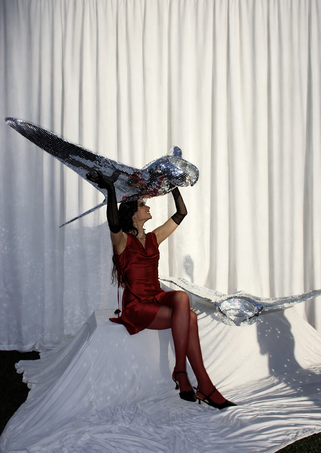 Woman in red dress and high heels sitting on a white surface holding a large, shiny, bird-shaped sculpture against a white curtain backdrop.