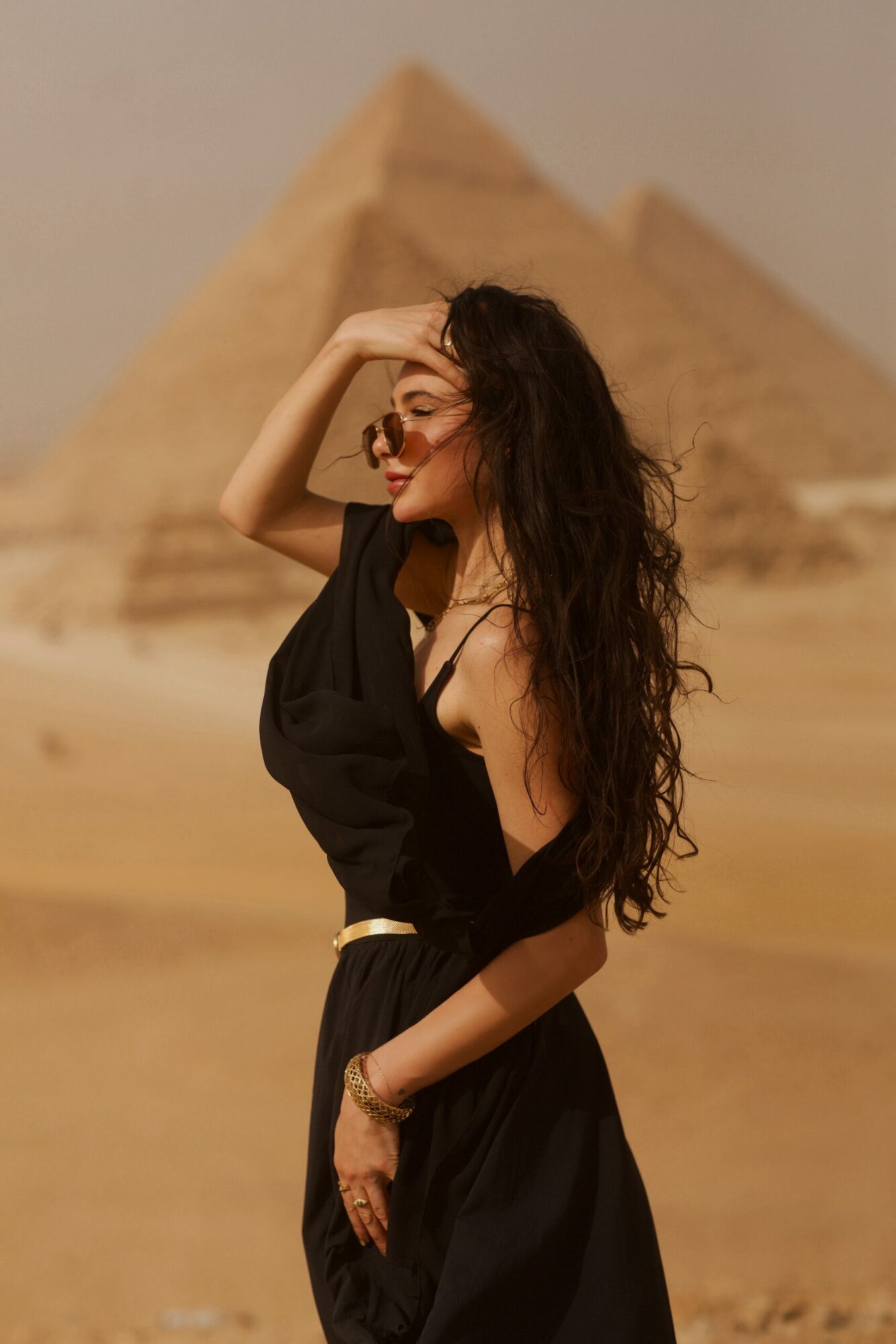 Woman with long curly hair and sunglasses standing in front of pyramid in desert.