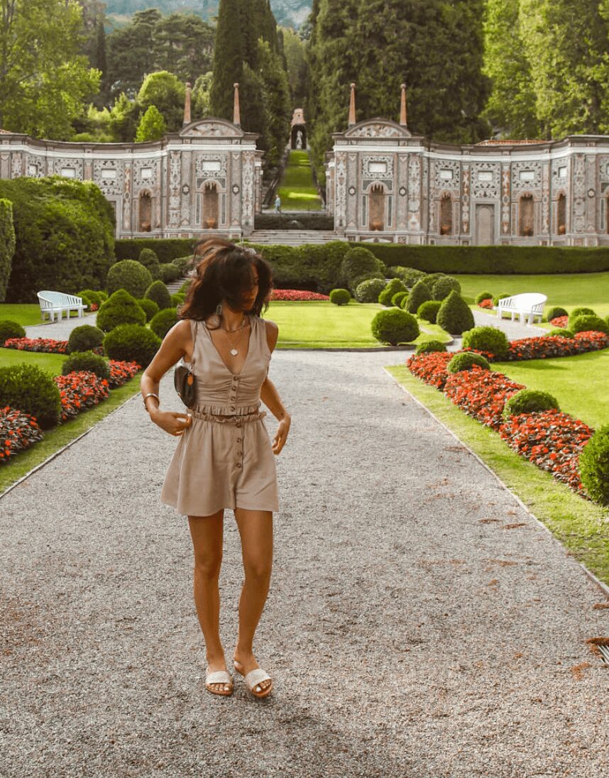 Woman walking on gravel path in a garden with trimmed bushes and ornate buildings in background.