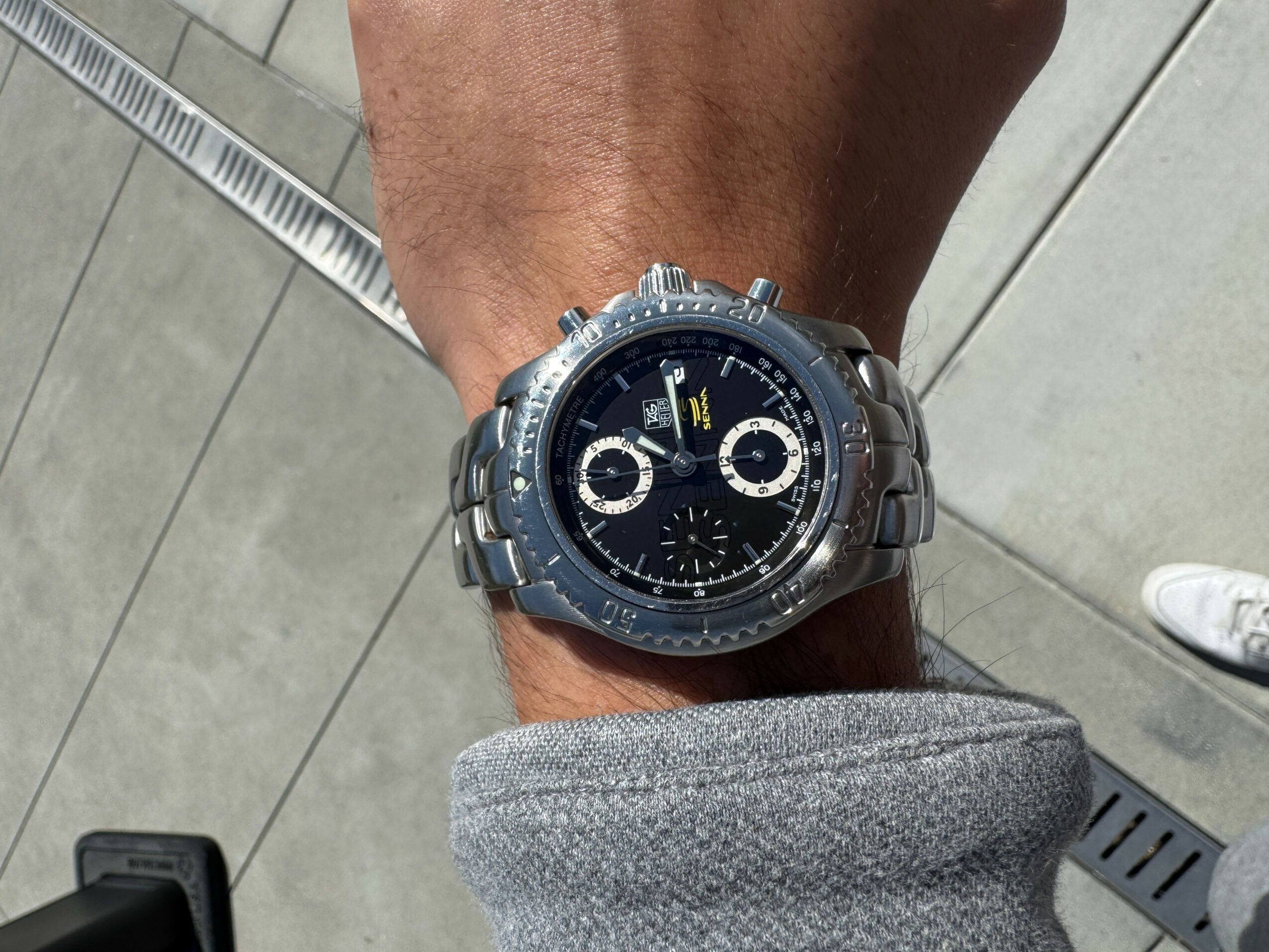 Person's wrist wearing a large silver watch with a black dial and multiple subdials, on a tiled floor.