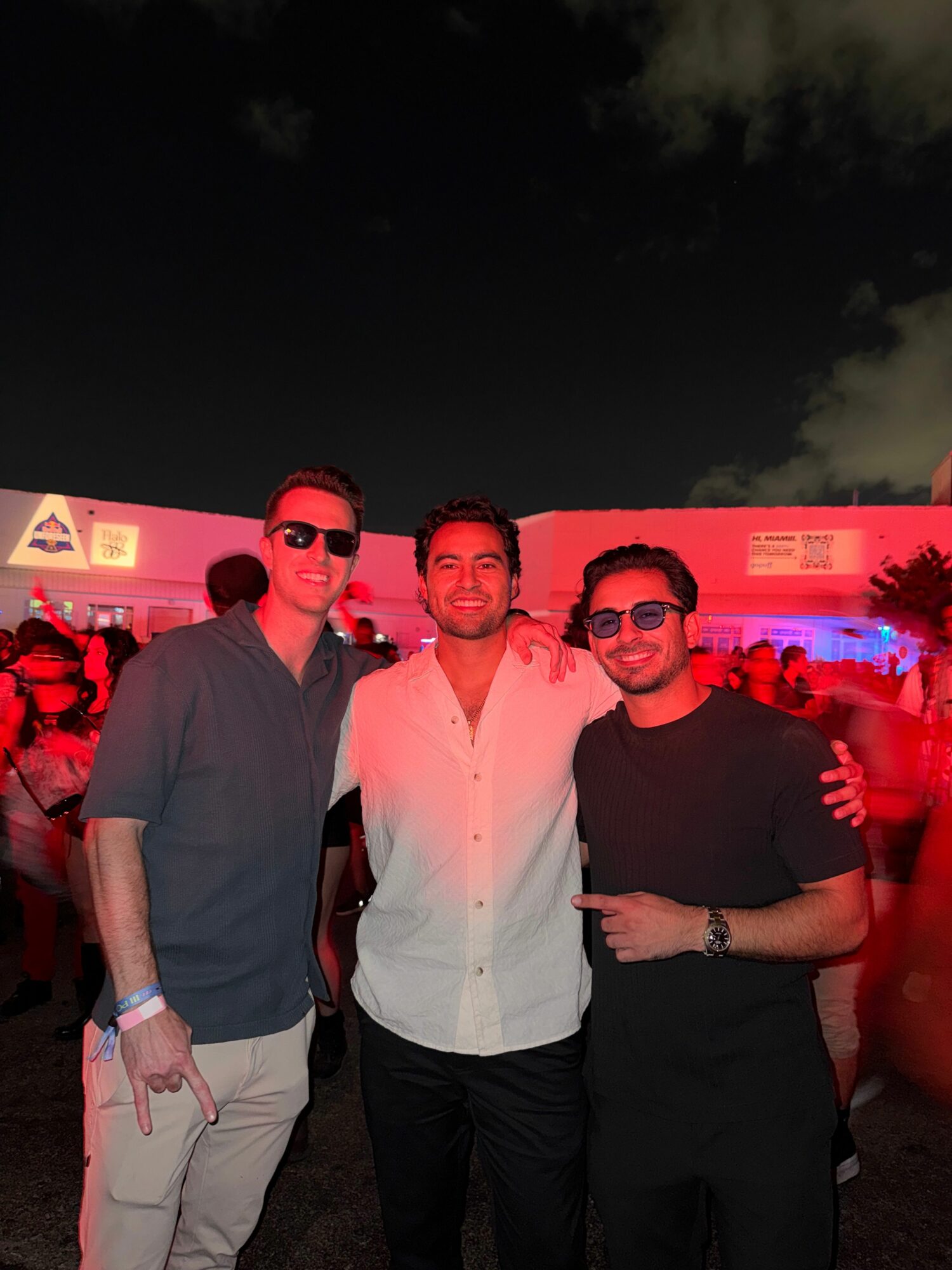 Three men standing outdoors at night, smiling, with a red-lit background and cloudy sky.