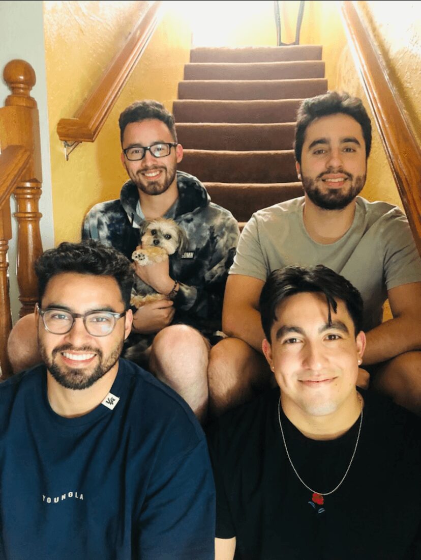 Four men sitting on stairs, smiling, with a small dog held by one person, staircase with wooden railing behind them.