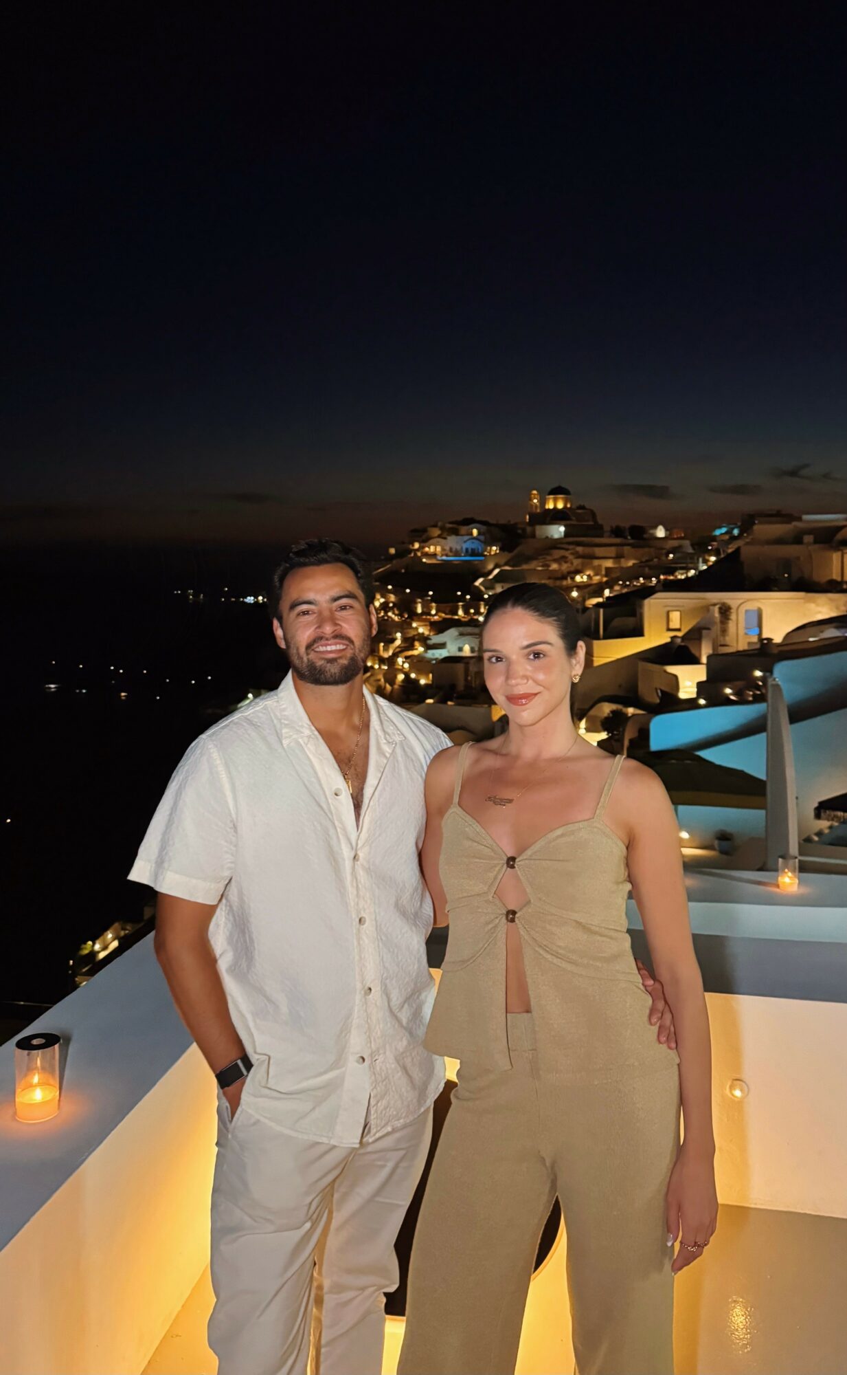 Two people stand on a rooftop at night with city lights and buildings in the background.