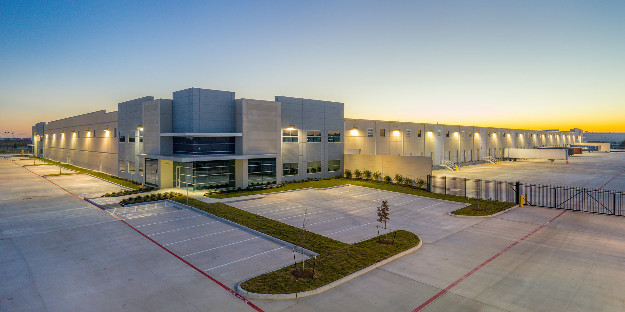 Large modern building with parking lot and landscaped areas, under a clear sky at sunset or sunrise.