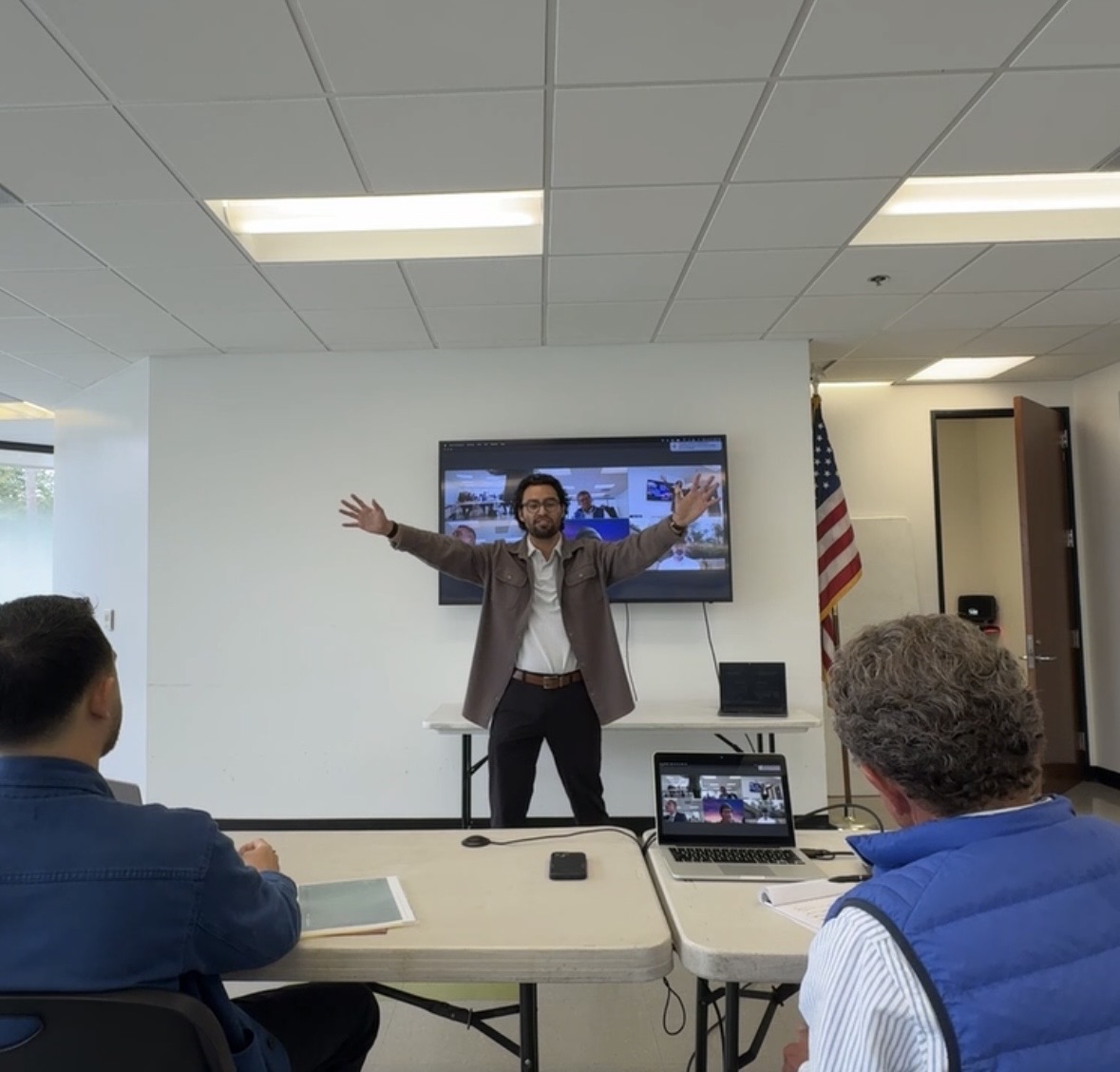 Man standing with arms outstretched in front of a screen, in a room with seated people and an American flag.