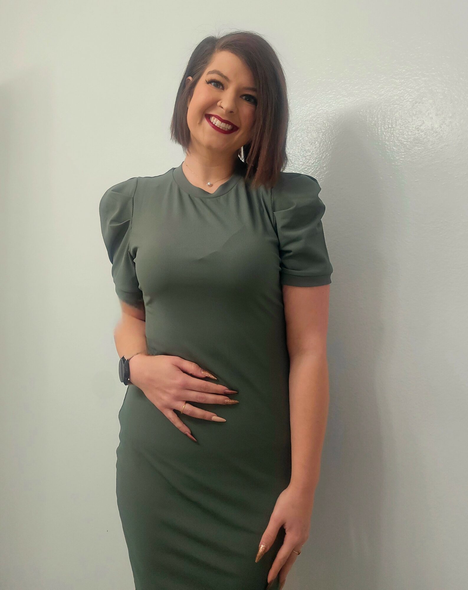 Woman with shoulder-length dark hair smiling, wearing a green dress with puffed sleeves, standing against a plain wall.