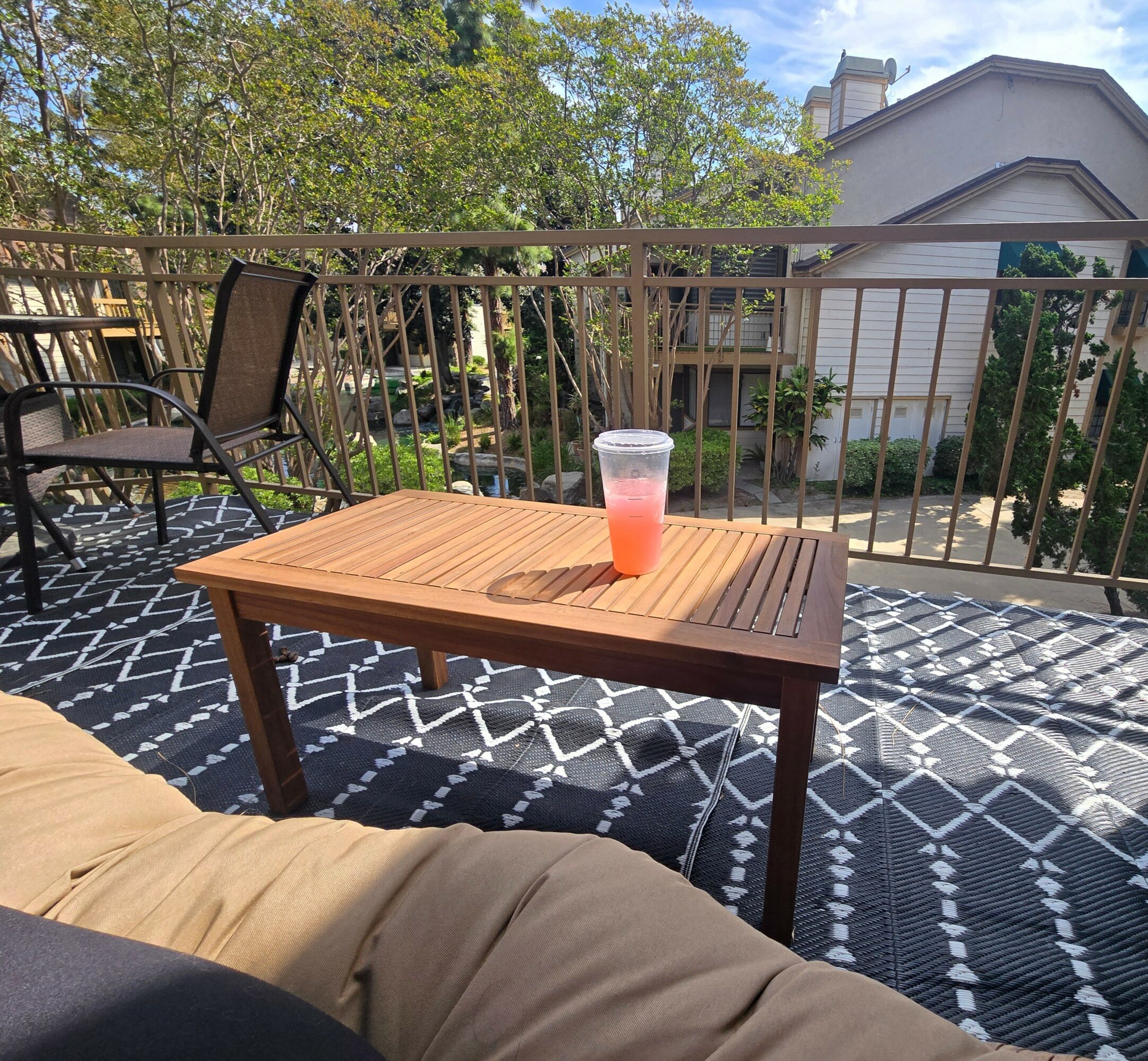 Wooden table with a pink drink in a clear cup on a patterned outdoor rug, balcony with railing, trees, and house in background.