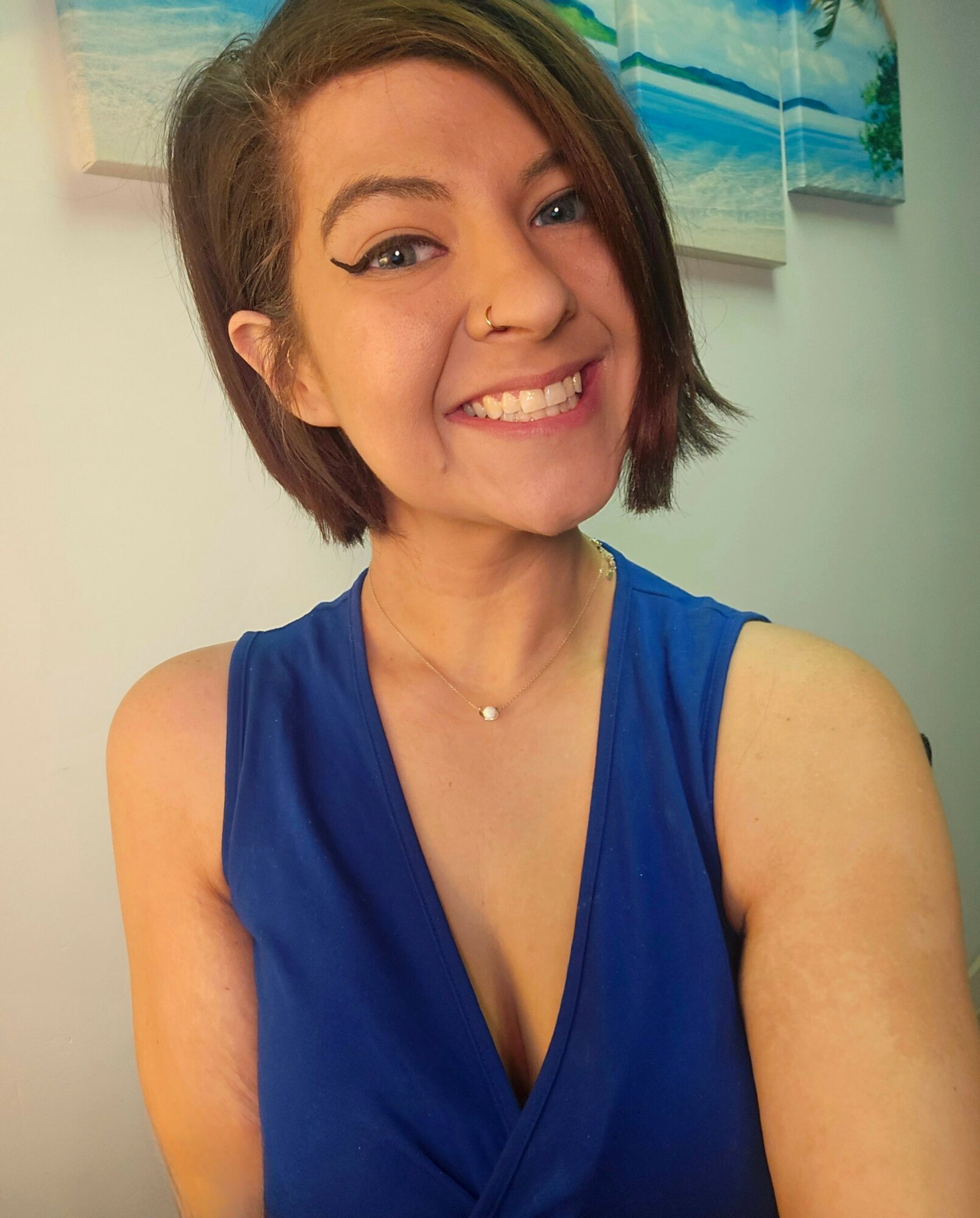 Young woman with short brown hair smiling, wearing a blue sleeveless top and a necklace, in front of a wall with artwork.