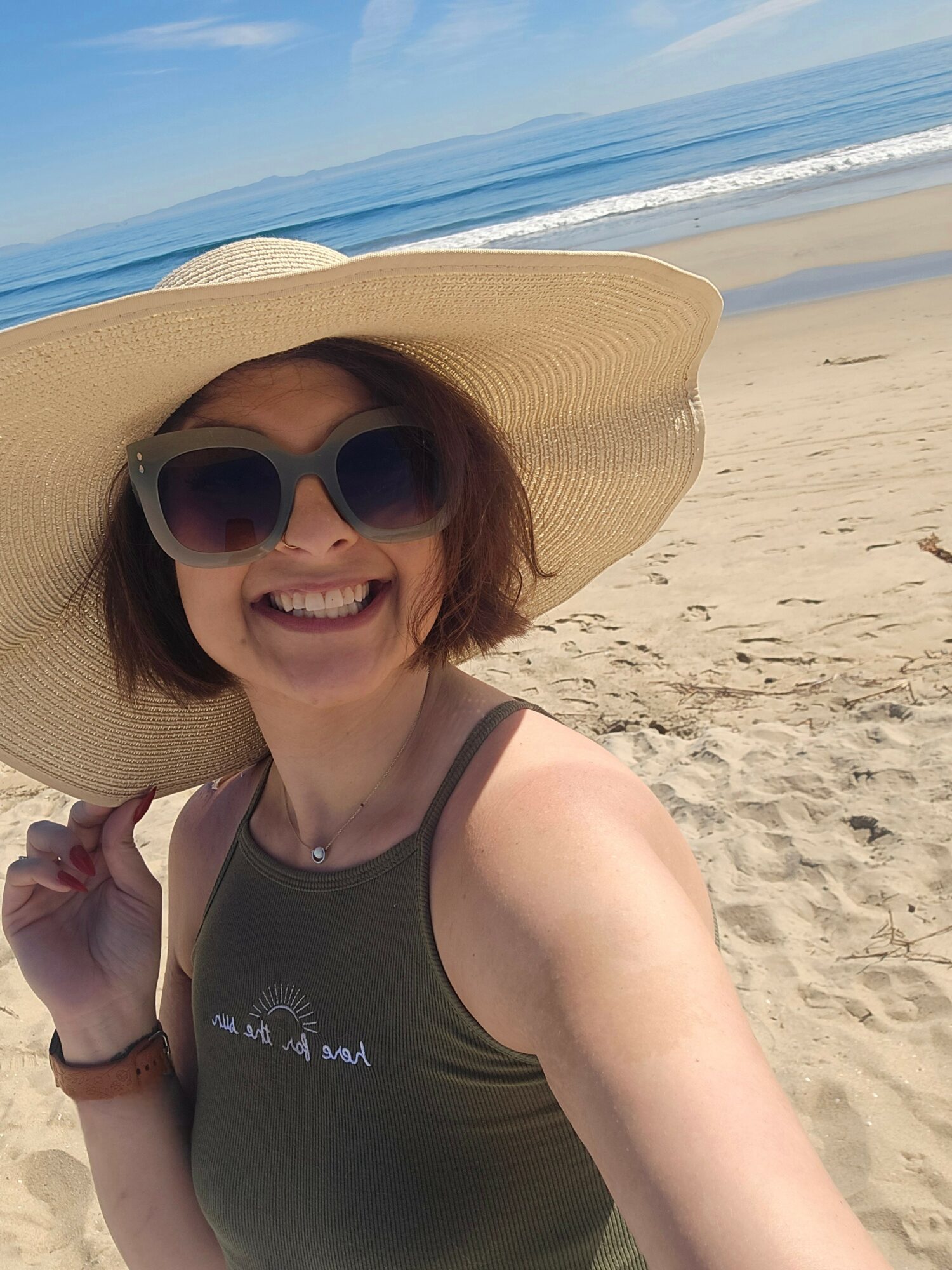 Woman smiling on beach wearing large sun hat and sunglasses, with ocean and sand in background.