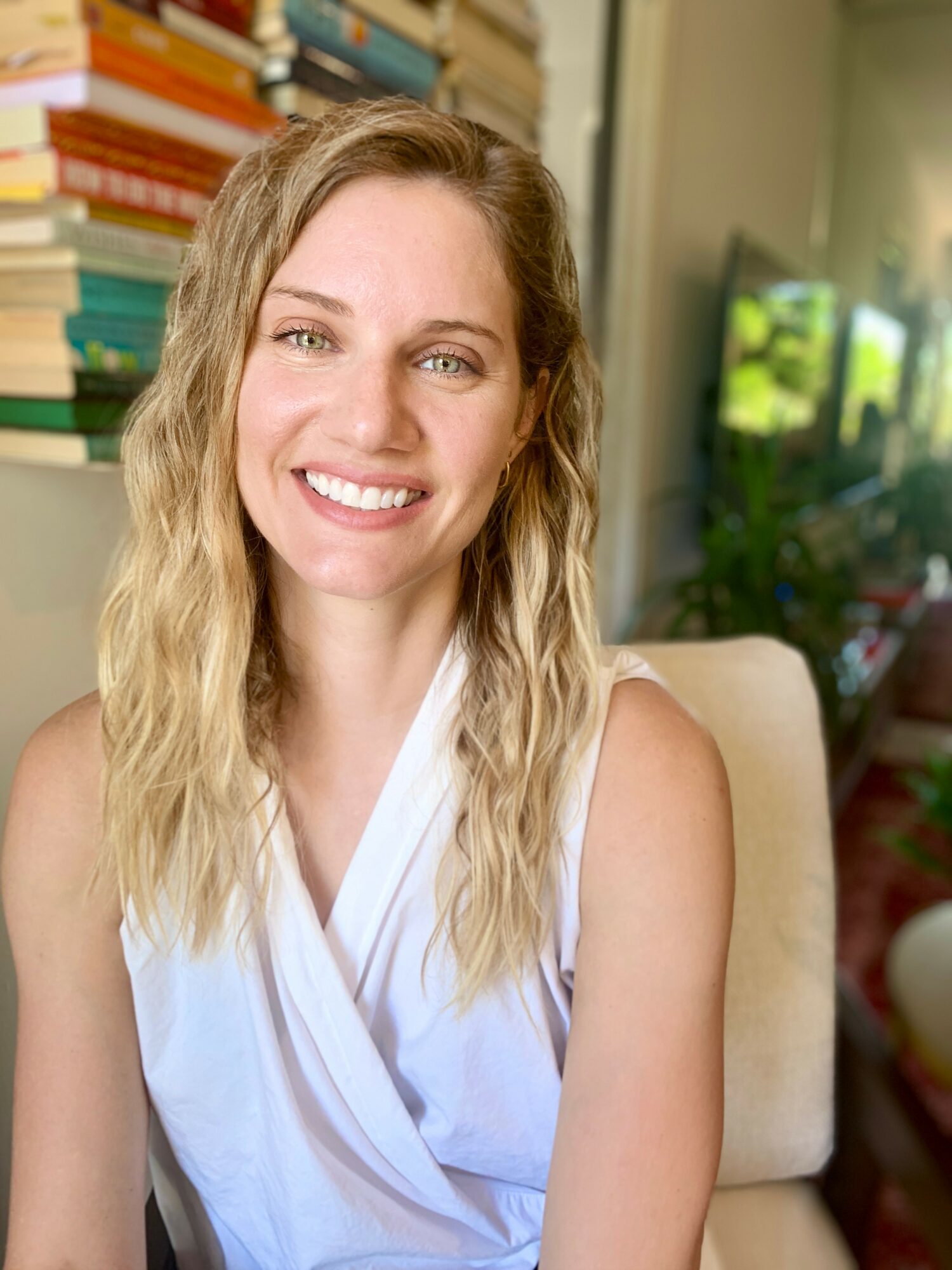 Young woman with wavy blonde hair smiling, sitting indoors with books and a TV in background.