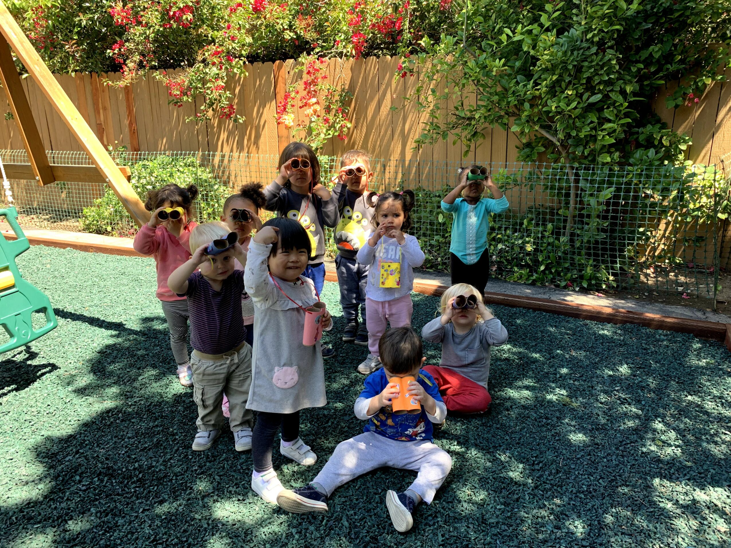 Group of children and an adult in a playground with green turf, wooden fence, and trees in background.