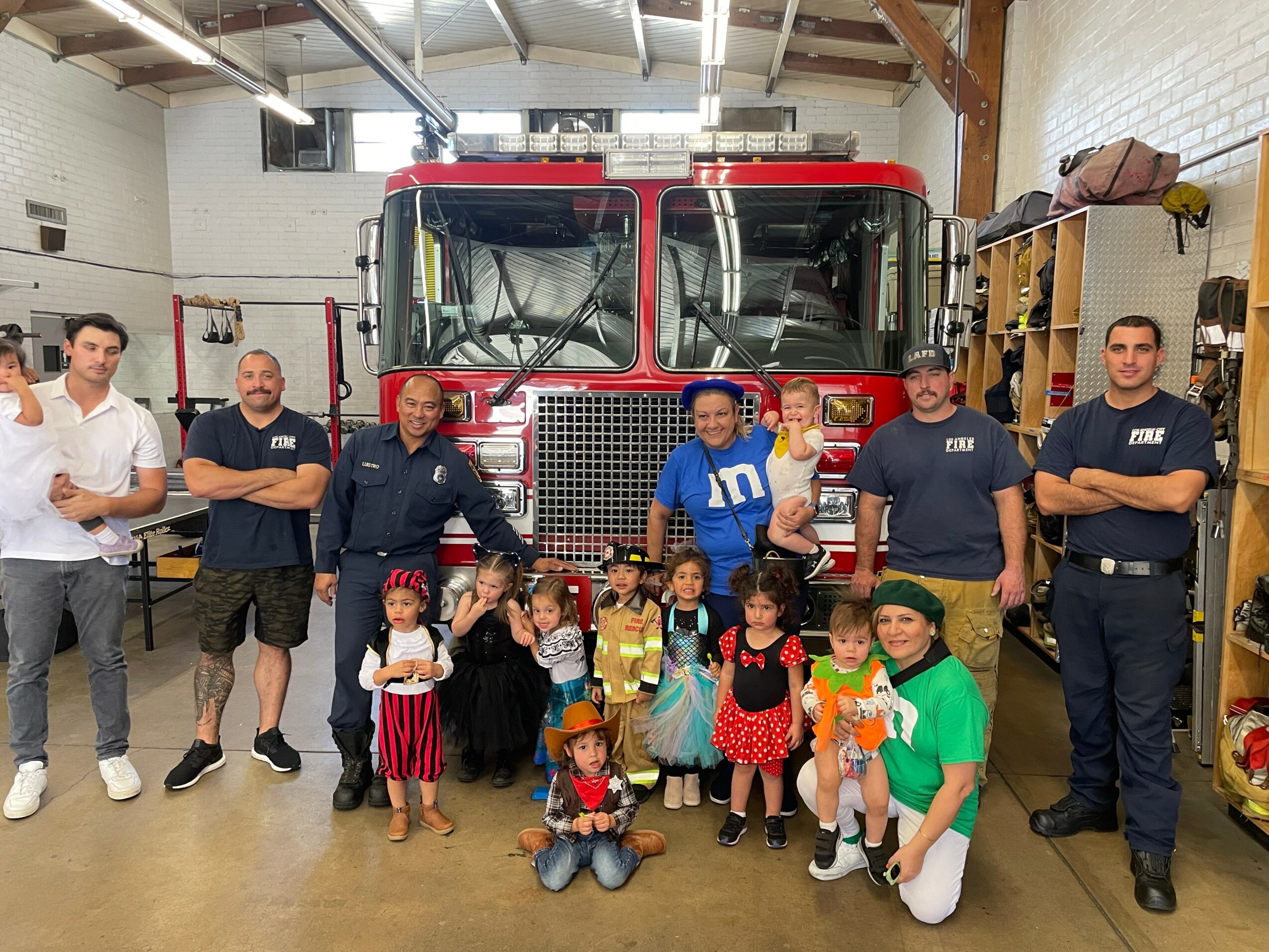 Group of children and adults standing in front of a fire truck inside a garage, some children holding toys.