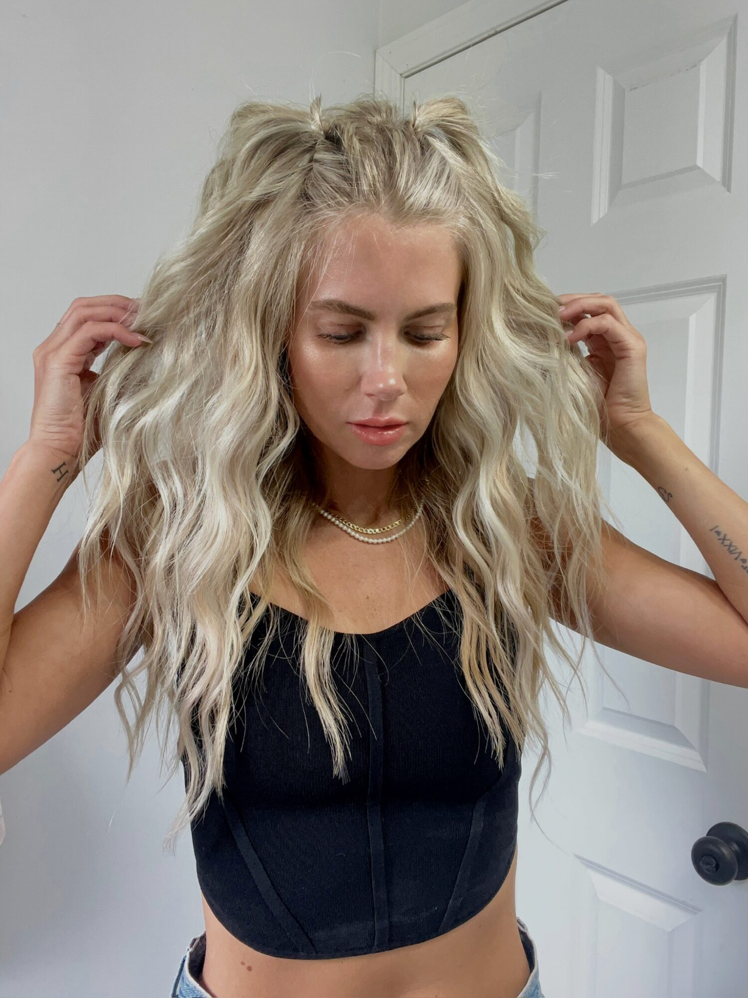 Woman with long, wavy blonde hair adjusting her hair, wearing a black top and a necklace, in front of a white door.