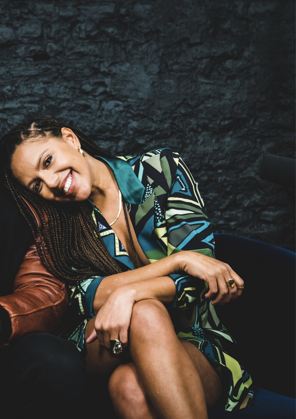 Smiling woman with braided hair sitting against a dark stone wall, wearing a colorful patterned jacket.