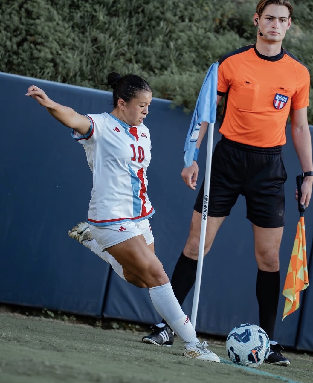 Female soccer player kicking a ball near sideline with assistant referee holding flag nearby.
