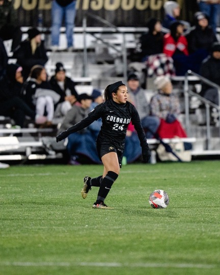Female soccer player in black uniform with number 24, running on field with ball, spectators in background.