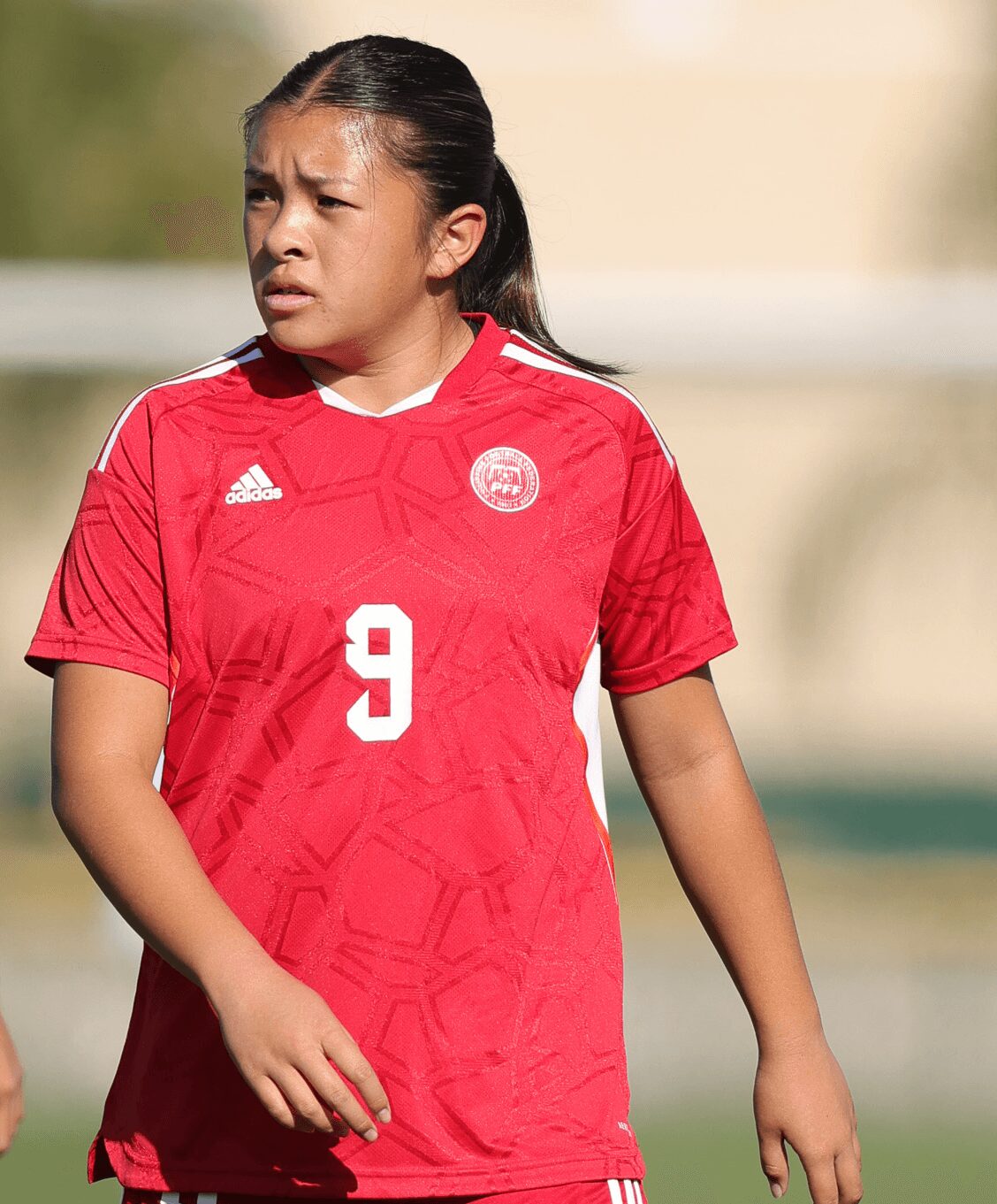 Young girl in red sports jersey with number 9, standing outdoors with a focused expression.