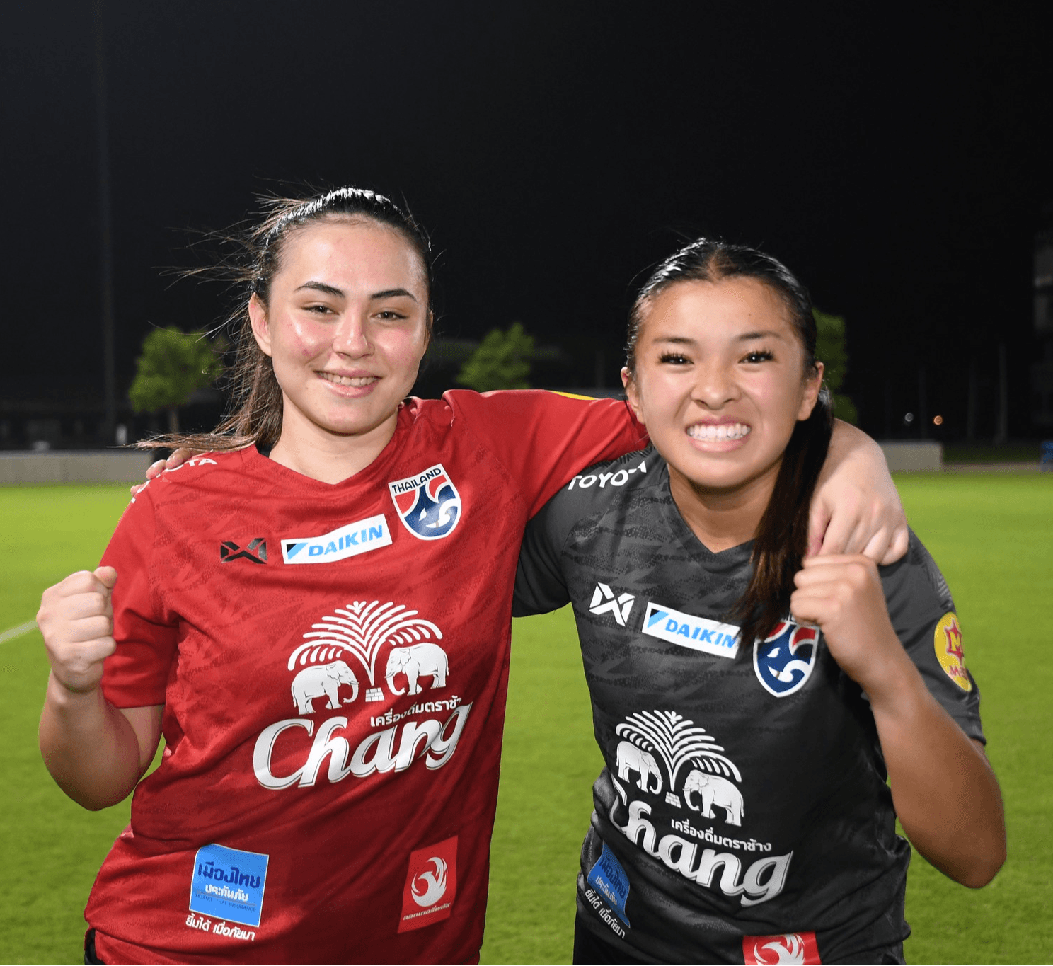 Two female soccer players smiling and celebrating on the field at night, wearing team jerseys and raising fists.