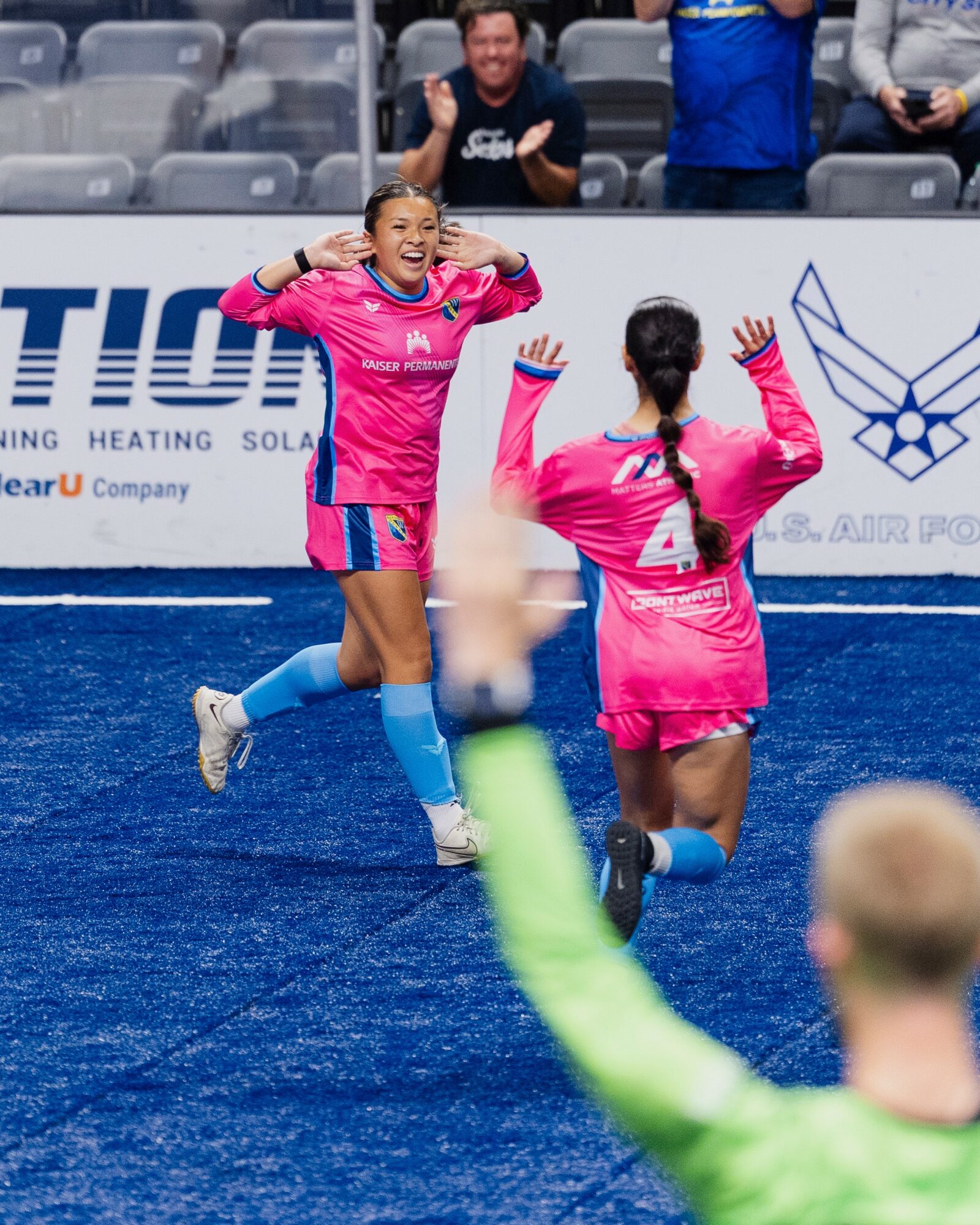 Two female soccer players in pink uniforms celebrate on the field, with one smiling and the other raising her hands.