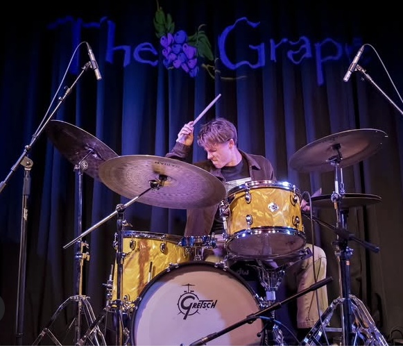 Young person playing yellow drum set on stage with dark backdrop and purple grape decoration.