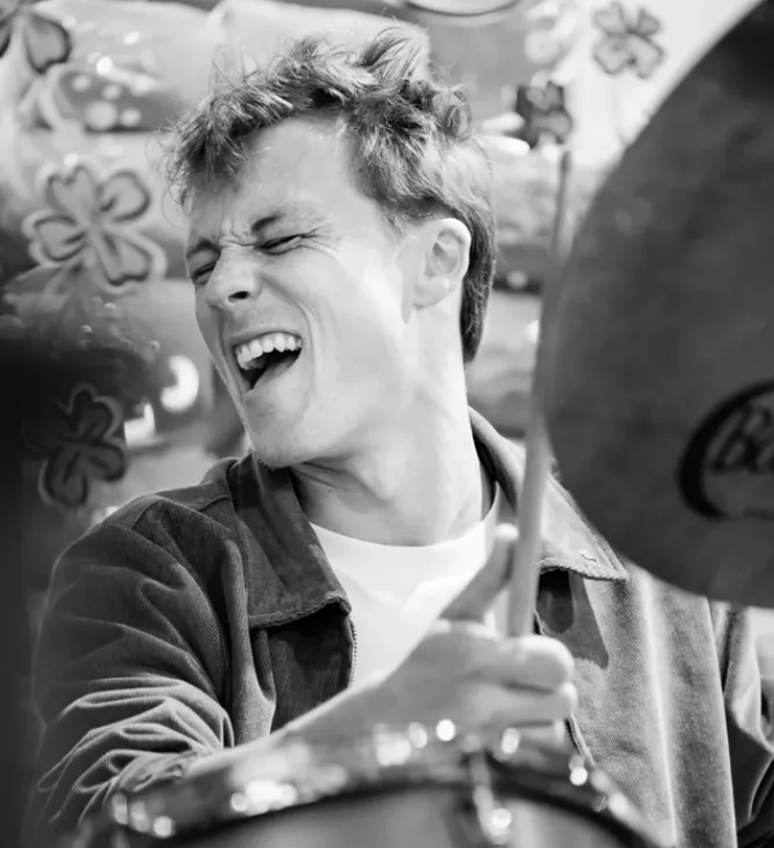 Person laughing with eyes closed, holding drumsticks, in front of a decorated background.