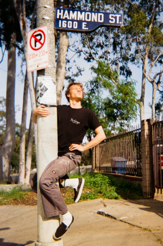 Person leaning against a pole on a sidewalk, with trees and a fence in the background.