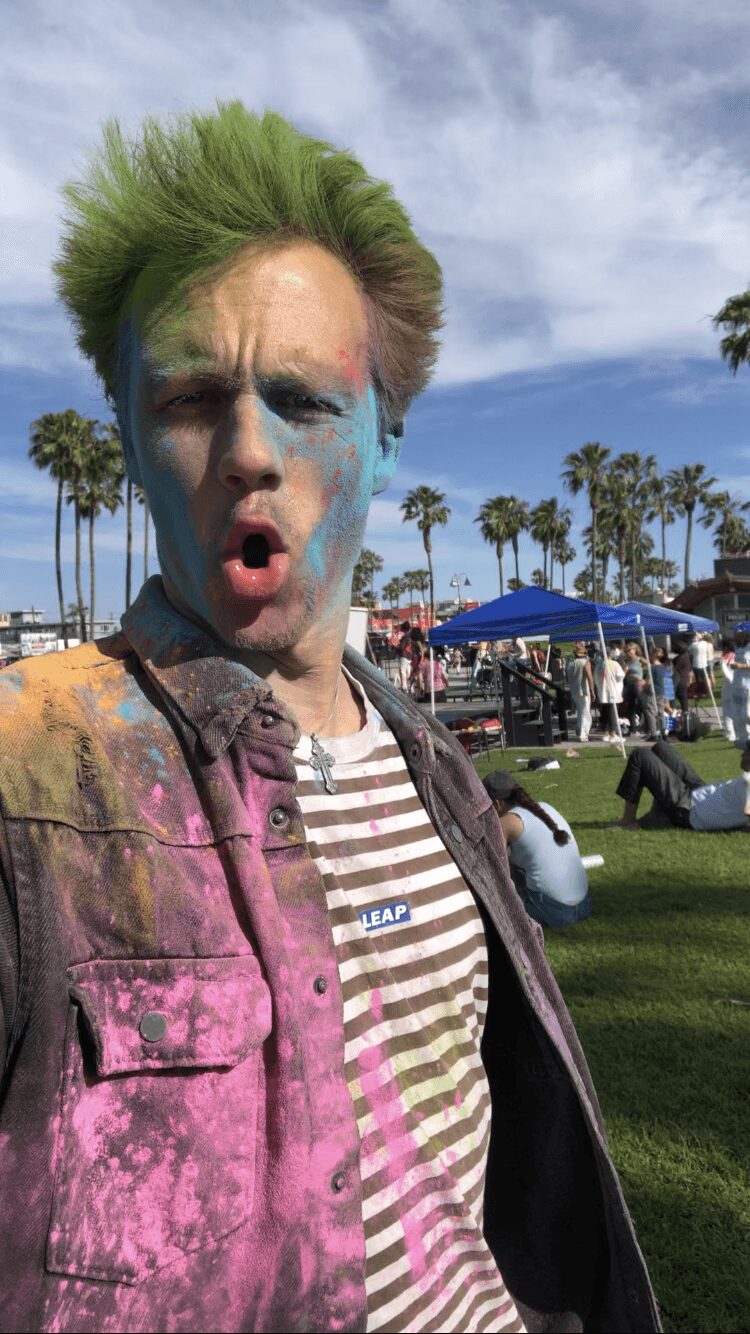 Person with green hair and face paint, wearing striped shirt and colorful jacket, outdoors with palm trees and tents.
