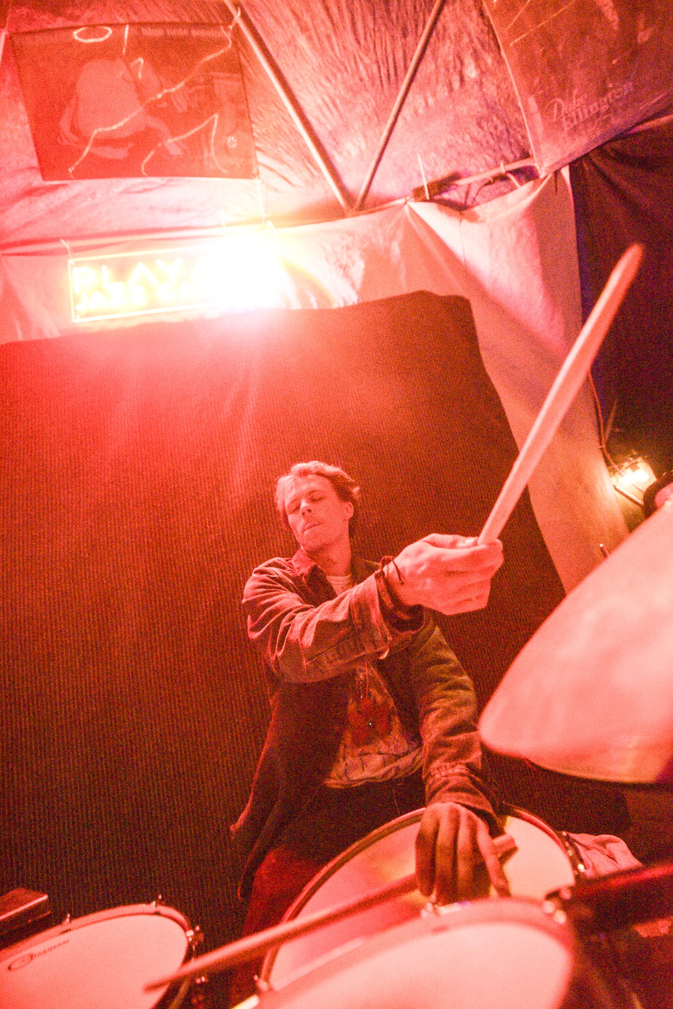 Person holding a drumstick on stage with drums, illuminated by red lighting, in a performance setting.