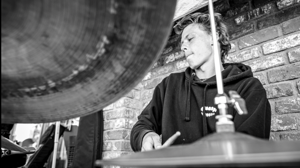 Young person playing drums against a brick wall, with a large cymbal in the foreground, in black and white.
