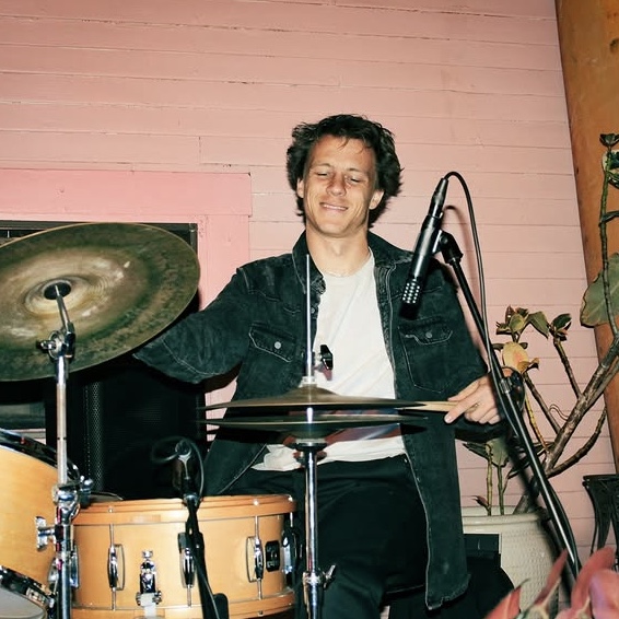 Person playing drums with a smile, wearing a dark jacket, in a room with pink wall and plants.