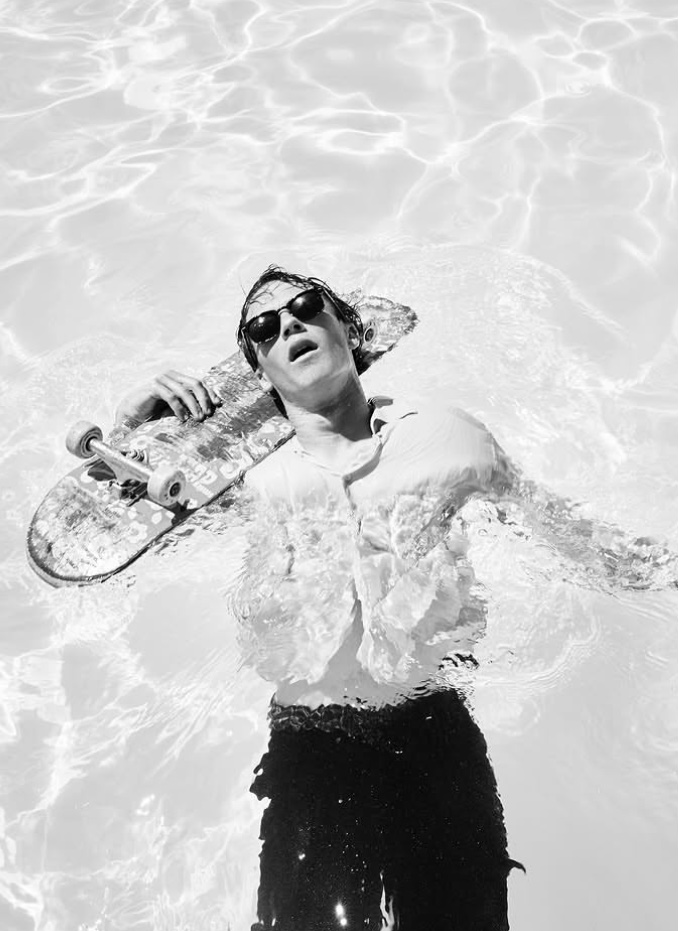 Person with sunglasses holding skateboard in water, smiling, wearing a white shirt and dark pants.