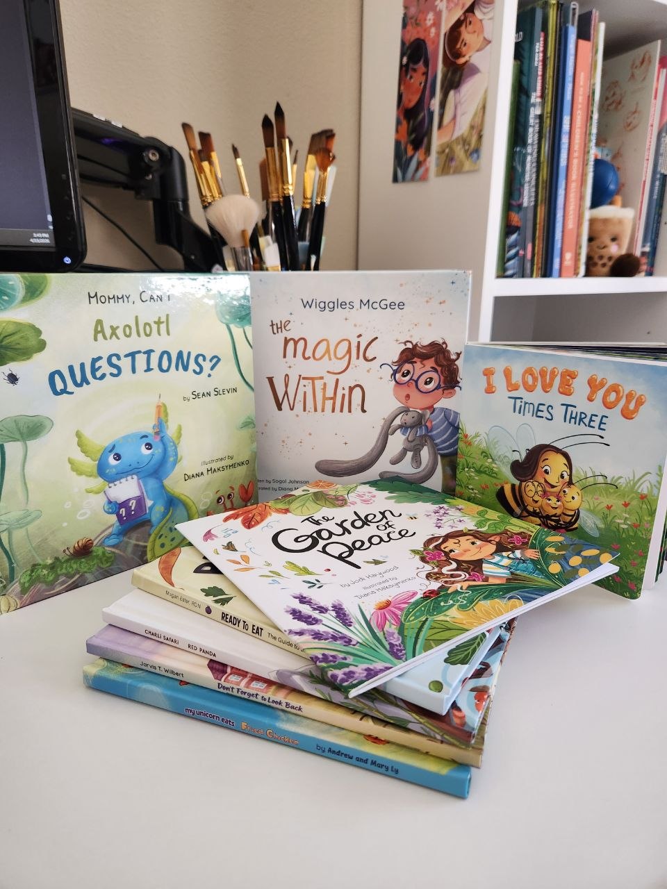 Stack of children's books on a white surface with colorful covers and illustrations, background includes art supplies and bookshelves.