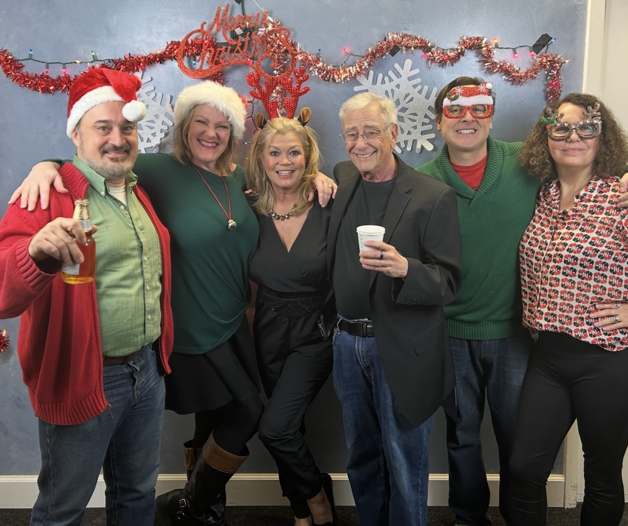 Six people wearing holiday accessories and clothing, standing in front of Christmas decorations, smiling for a group photo.