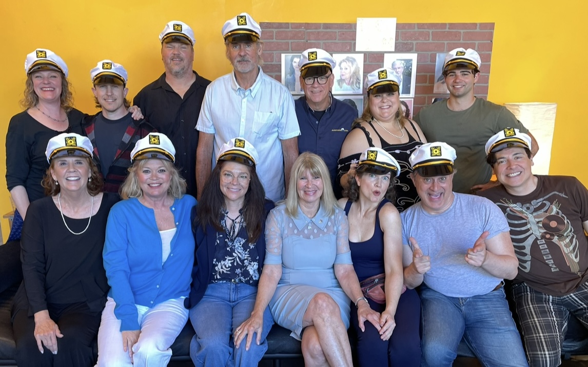 Group of 13 people wearing hats, sitting and standing in front of a yellow wall.