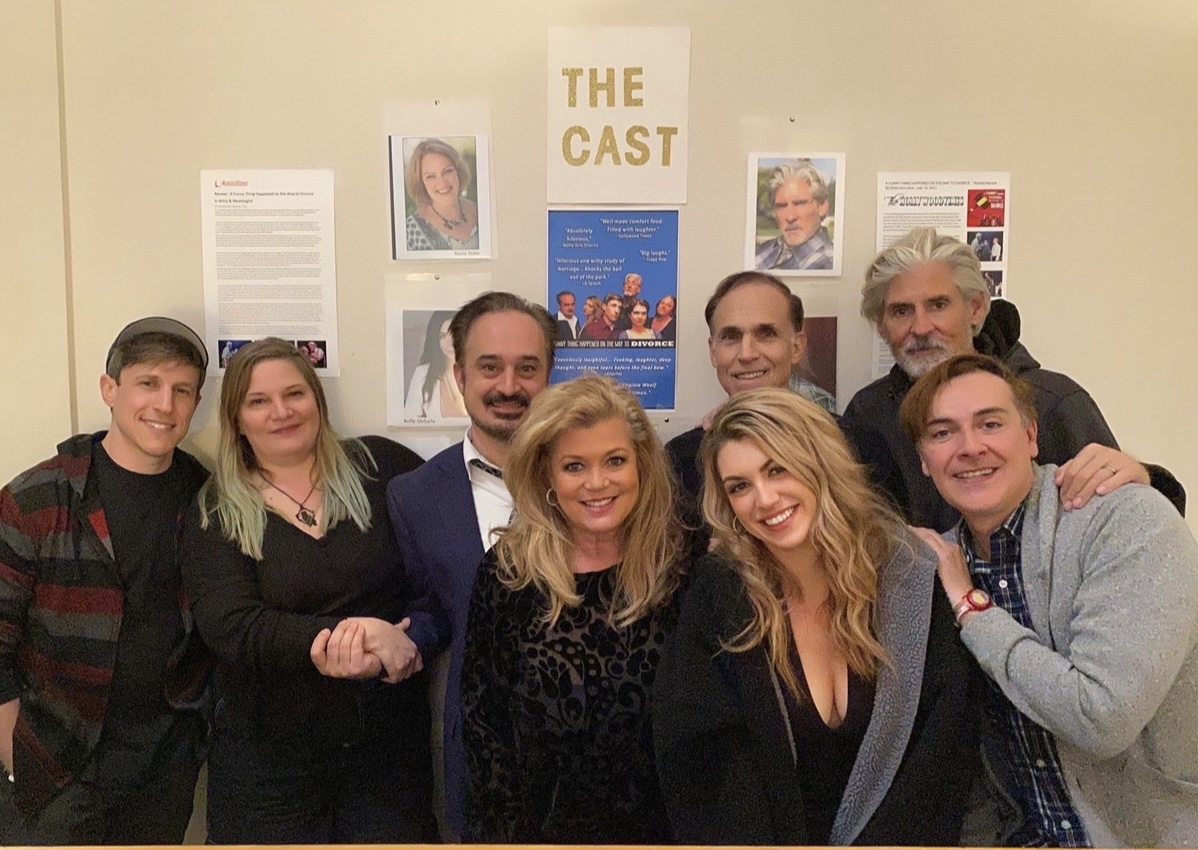 Group of eight people smiling and posing together indoors, behind a wall with posters and photos.