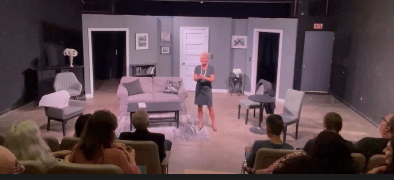 Stage with a woman speaking, surrounded by chairs and audience members, in a theater or studio setting.