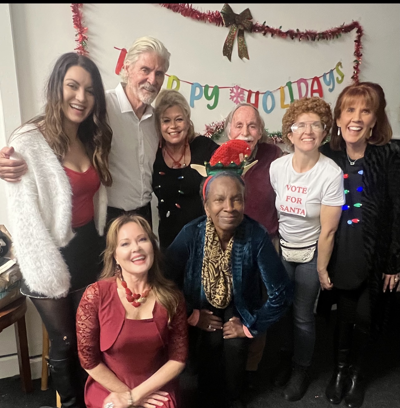 Group of eight people celebrating Christmas, standing in front of a decorated wall with a 'Happy Holidays' banner.