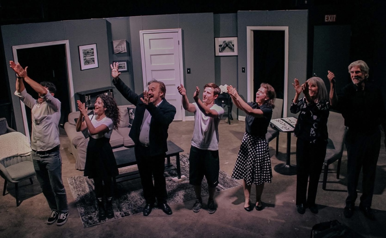 Group of eight people holding hands and smiling on a stage set resembling a living room.