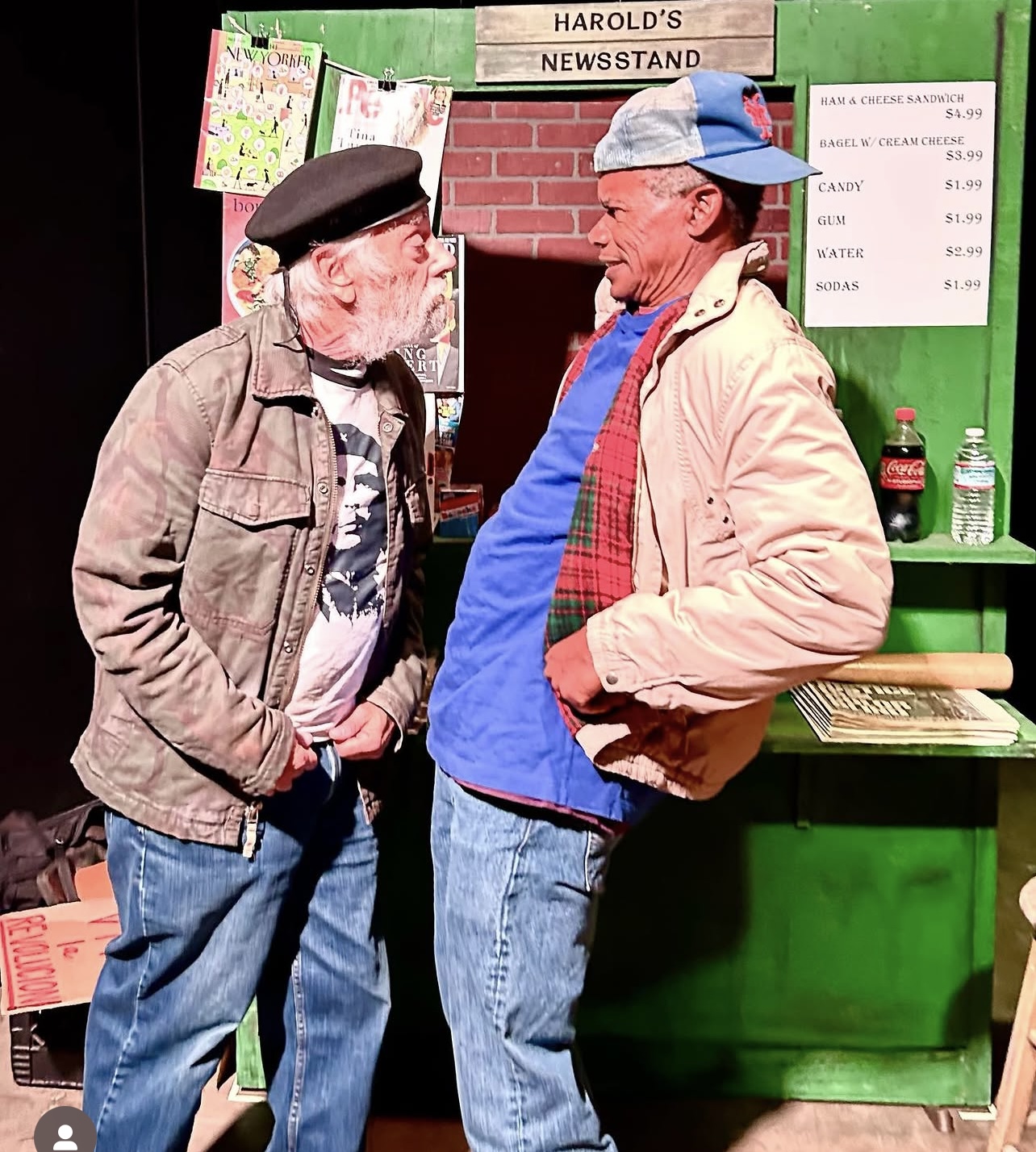 Two men with beards and hats talking in front of a green newsstand with signs and water bottles.